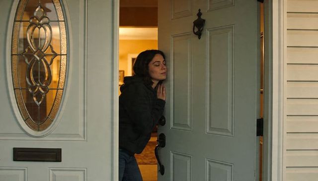 Woman leaning at front door with oval glass closing eyes touching chest in reflection