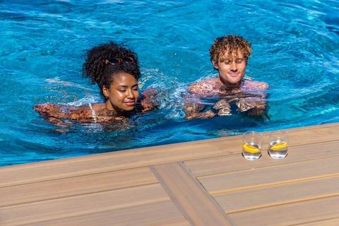 Sunlit resort pool with diverse couple swimming side-by-side near wooden deck and lemon water