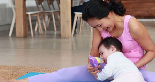A young Asian woman enjoys a bonding moment with her baby during a break in her yoga routine, with copy space. She smiles gently as the infant engages with a colorful toy, highlighting the balance of motherhood and personal wellness.