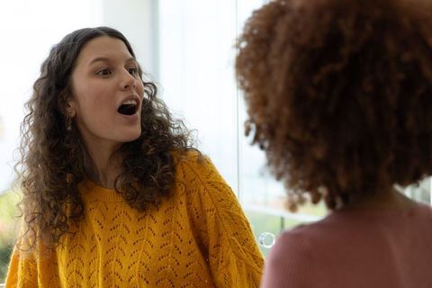 Diverse female friends laughing and blowing soap bubble by window, mustard sweater