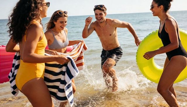 Friends splashing and laughing on sunny beach with inflatable ring and colorful towels
