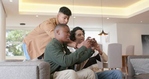 Three friends sharing smartphone and playing mobile game on living room couch