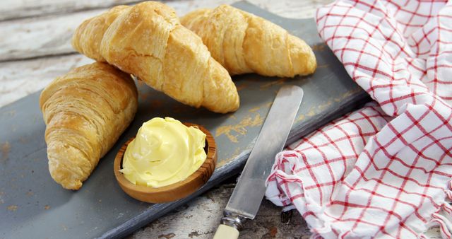 Freshly baked croissants are paired with creamy butter on a rustic kitchen table, with copy space. A red and white checkered napkin adds a cozy, homely touch to the delightful breakfast setting.