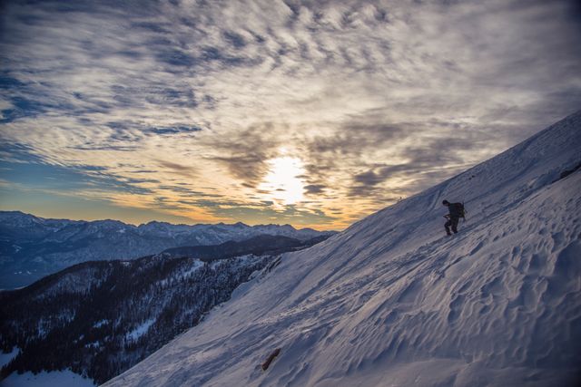 Picture of a climber ascending a snow-covered mountain slope during sunset, casting a dramatic sky with scattered clouds. The scene captures the intensity and solitude of mountaineering, ideal for use in themes related to adventure, extreme sports, motivation, and natural beauty.