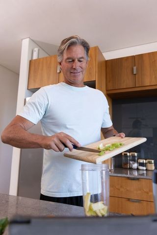 Senior man preparing green apple smoothie at kitchen counter in cozy modern home