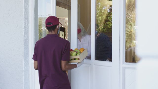 A woman accepts a box of fruits and vegetables from a delivery person at the entrance of her house, both adhering to COVID-19 safety protocols like wearing face masks and gloves. Perfect for illustrating stories on safety, contactless deliveries, sustainable grocery service, pandemic precautions, and logistical advancements during challenging times. Suitable for content about lifestyle changes brought by COVID-19.