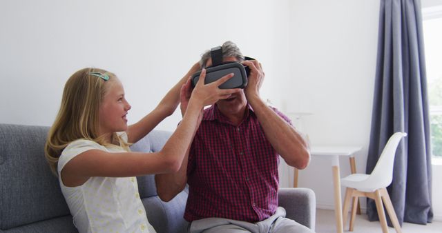 Granddaughter helping grandfather put on a virtual reality headset in the living room. This image can be used for promoting family technology products, illustrating bonding moments between different generations, or highlighting the playful use of modern technology in everyday life.