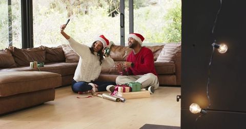 Diverse couple wearing Santa hats taking selfie while wrapping Christmas gifts at home