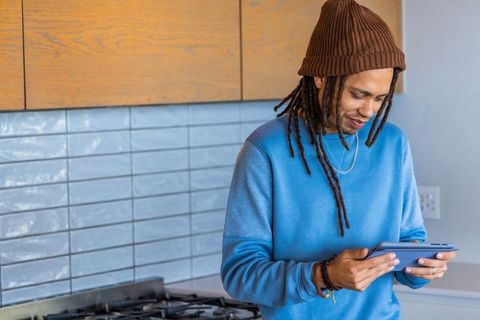African american man wearing beanie holding tablet and looking in minimalist kitchen