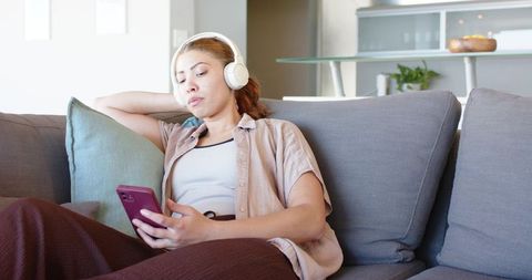 Young woman relaxing on sofa with headphones and smartphone, modern living room lifestyle