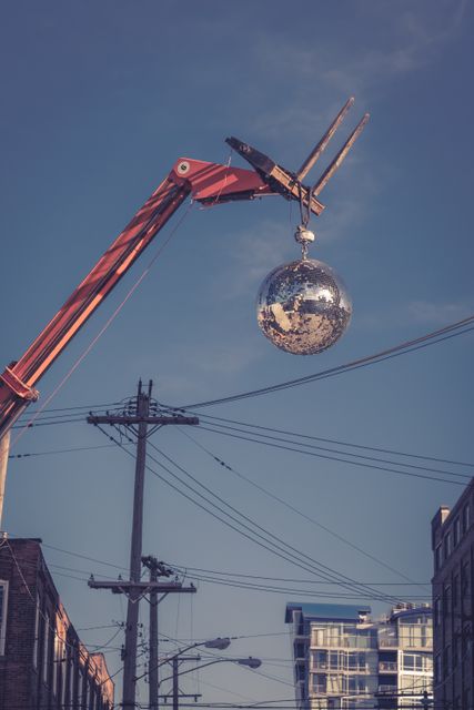 Vintage crane lifting a disco ball in an urban area. The cityscape includes utility poles and buildings, combining elements of old machinery with modern architecture. Useful for illustrating urban industrial scenes, construction work, and creative installations in public spaces.