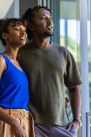 African american couple standing by sunlit glass door looking out together on balcony