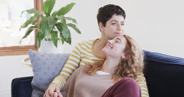 Happy caucasian lesbian couple sitting,embracing and holding hands on sofa in sunny house. Togetherness, relationship, lifestyle, relaxation,domestic life, unaltered.