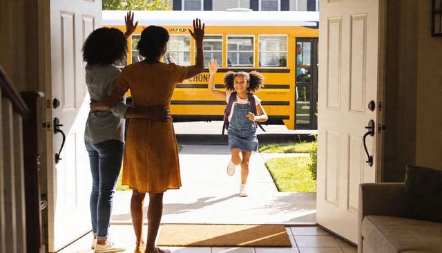 Schoolgirl running home waving to joyful guardians at open front door with yellow bus