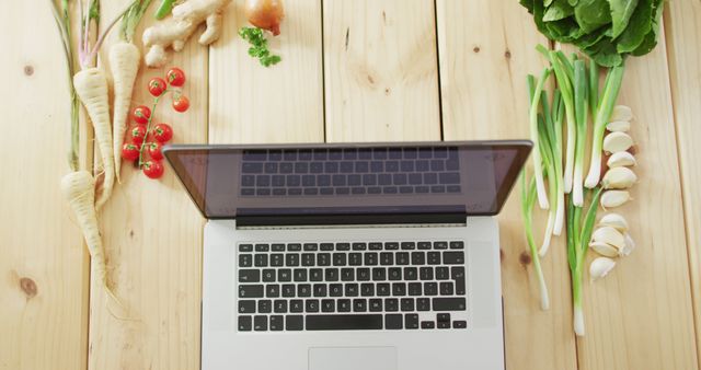 Top view of laptop on wooden table surrounded by fresh vegetables like ginger, cherry tomatoes, spring onions, parsnips, and garlic. Perfect for themes around balancing work and healthy eating, nutritional blogs, work-life balance, culinary technology, and remote work setups.