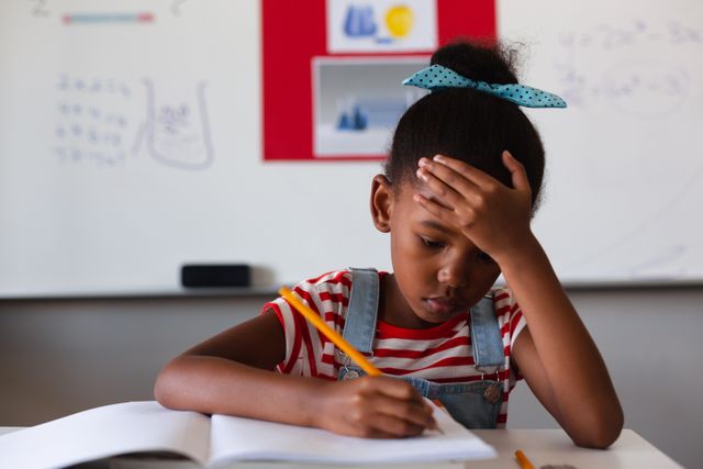 African american elementary schoolgirl with head in hand writing on book at desk in classroom. unaltered, education, learning, studying, concentration and school concept.