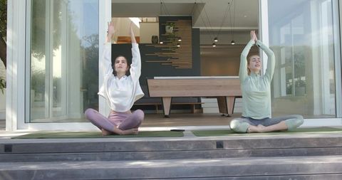 Two women practicing yoga and stretching on sunlit deck outside modern serene glass house