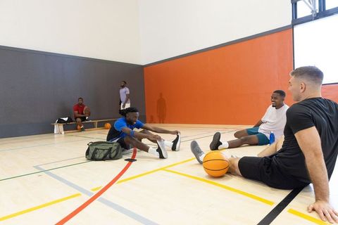 Diverse teammates stretching and laughing on indoor basketball court during warmup