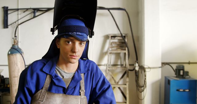 Female welder in blue overalls and protective gear working in an industrial workshop environment. She is using safety equipment including a welding helmet and gloves. The workshop features typical tools and machinery associated with metal fabrication. This image can be used in articles and advertisements focused on industrial jobs, women's empowerment in the workplace, and occupational safety.