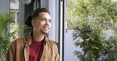 Mid-adult man leaning by sliding door with houseplant smiling and looking outside