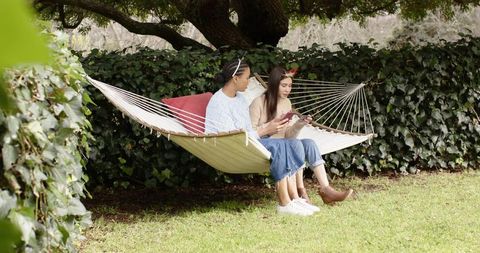 Two friends relaxing on hammock sharing smartphone and laughing in leafy backyard garden