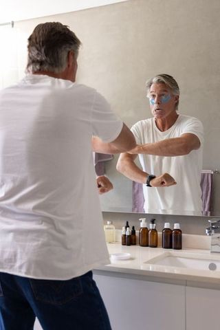 Mature man applying under-eye patches while checking mirror for morning skincare routine
