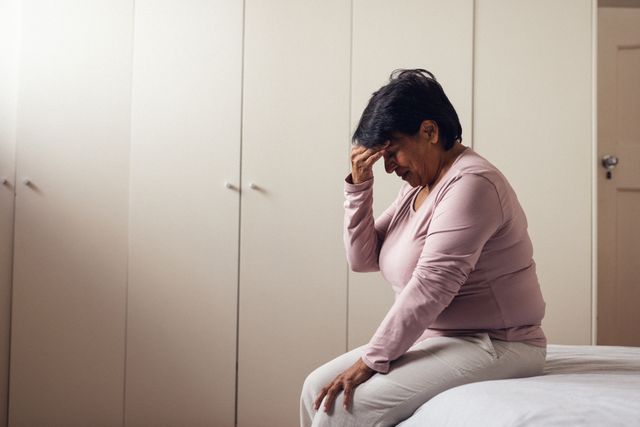Side view of biracial mature woman with headache sitting on bed against closets at home, copy space. Short hair, pain, unaltered, healthcare, sickness and retirement concept.