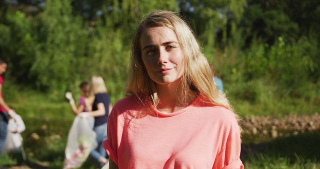 The young woman in a pink shirt participates in an environmental cleanup effort in a lush, green park. She appears optimistic and proud of her contributions while other volunteers in the background gather litter in plastic bags. This image is ideal for content related to community volunteering, environmental conservation, and sustainability initiatives.