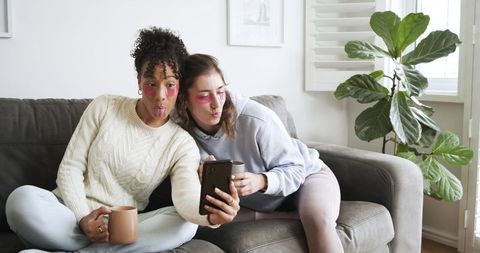 Diverse friends wearing pink under-eye patches taking cozy couch selfie while holding mugs
