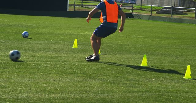 Soccer player wearing orange training vest practicing dribbling through cones on green football field. Great for content related to sports training, fitness routines, soccer tutorials, and athletic development.
