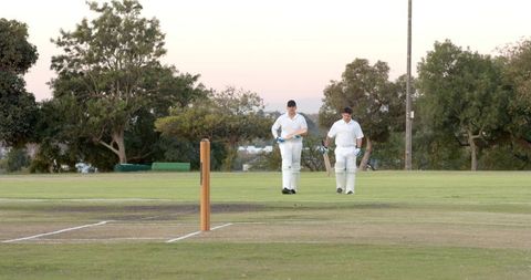 Two cricketers walking on pitch in white kit carrying bats returning to crease at sunset
