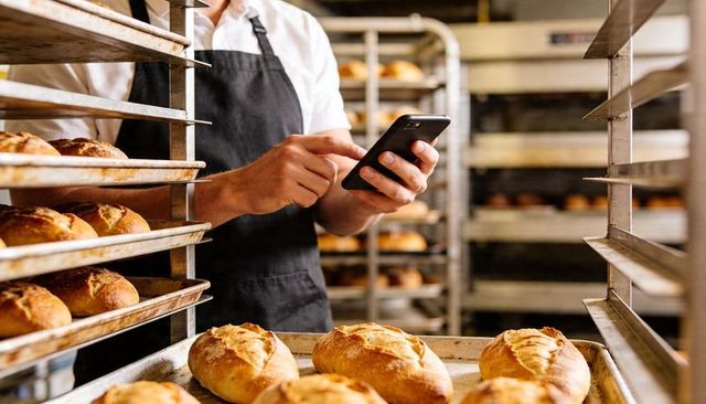 Baker Checking Smartphone Among Fresh Artisan Baguettes and Steel Racks