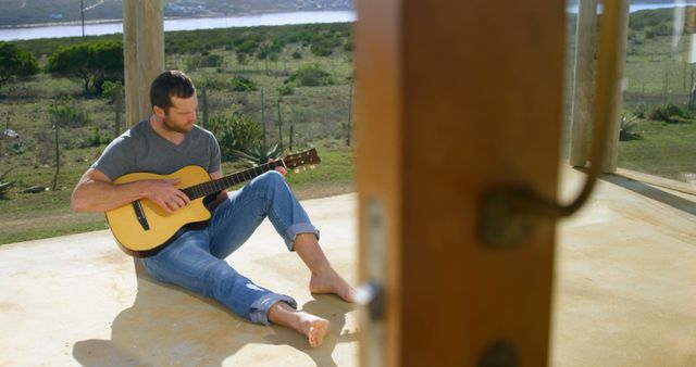 Middle-aged man sitting on an open porch playing an acoustic guitar while enjoying the serene rural surroundings. Barefoot and casually dressed, he appears to be in a moment of mindful relaxation, embracing nature. Great for concepts related to leisure activities, mindfulness, music, simplicity, rustic living, and stress relief.
