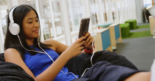 Young biracial woman enjoys music on her phone, with copy space. She's relaxing in a casual indoor setting, at home or a lounge area.