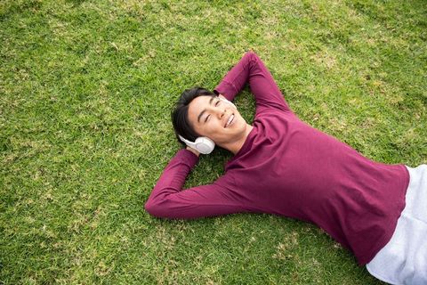Asian man lying on grass listening to music with headphones, relaxing outdoors