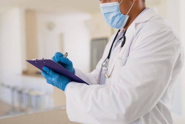 Hispanic male doctor wearing face mask and gloves, writing notes on a clipboard. Ideal for use in healthcare, medical, and pandemic-related content. Can be used to illustrate safety measures, professional healthcare services, and the importance of protective gear during the COVID-19 pandemic.