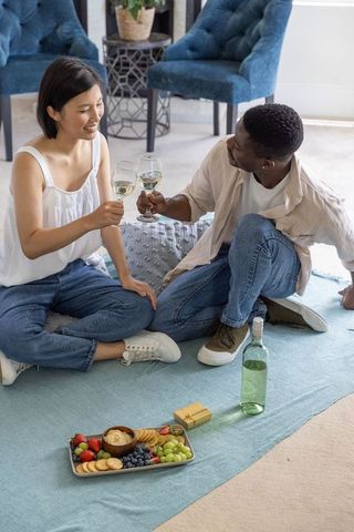 Multicultural couple enjoying cozy at-home picnic, clinking wine glasses, snack platter