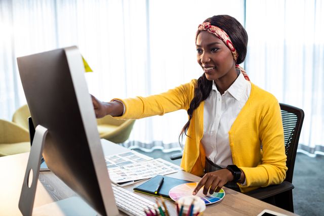 Businesswoman in a yellow sweater working on a computer in a modern office. She is pointing at the screen, indicating active engagement with her work. Ideal for use in articles or advertisements related to business, technology, office environments, professional women, and creative industries.