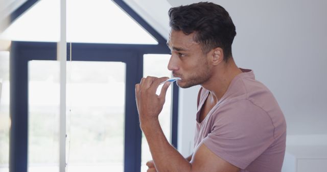 Young man standing in modern, minimalistic bathroom brushing teeth and focusing on oral hygiene. Image portrays a clean, fresh start to the day, ideal for advertisements on dental care, health blogs, and self-care promotions. Could be used in wellness websites, personal care guides, or morning routine content.