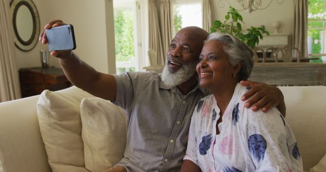 Happy senior couple taking a selfie on a cozy sofa in the living room. Ideal for representing themes of family, togetherness, happiness in retirement, and capturing memorable moments. Perfect for use in advertisements for retirement communities, family-oriented products, and home decor. Shows a joyful and content lifestyle indoors.