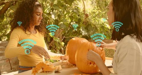 Two women carving pumpkins outdoors collaborating on diy autumn crafts with wifi icons