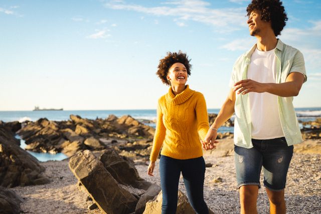 Young couple enjoying a walk on a rocky beach, holding hands and smiling. Ideal for use in lifestyle, travel, and relationship-themed projects. Perfect for promoting romantic getaways, outdoor activities, and healthy living.