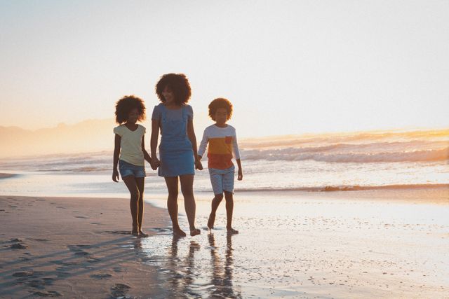 African American mother walking with her son and daughter on the beach at sunset. Ideal for themes of family bonding, summer vacations, outdoor activities, and relaxation by the sea. Perfect for travel brochures, family-oriented advertisements, and lifestyle blogs.
