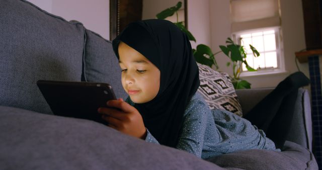A young girl of Middle Eastern ethnicity is focused on a smartphone while relaxing on a couch, with copy space. Her engagement with the device suggests a blend of technology and leisure in modern childhood.