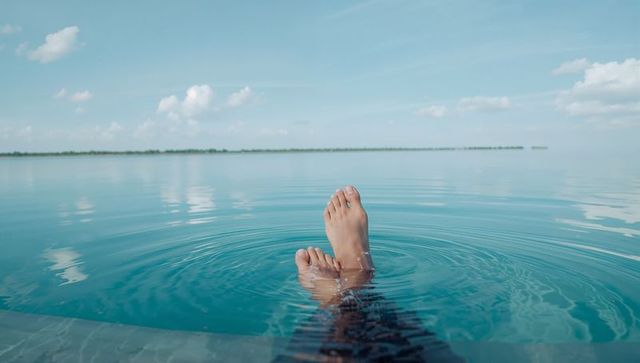 Relaxing bare feet in turquoise infinity pool with calm horizon and cloud reflections
