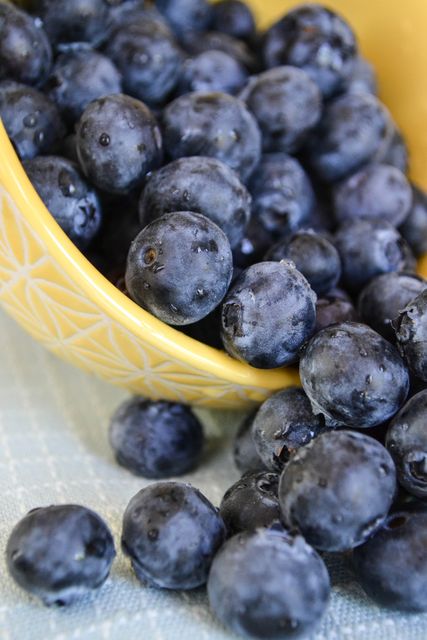 Close-up image of fresh blueberries spilling out of a patterned yellow bowl onto a cloth. Ideal for use in food blogs, healthy eating promotions, organic produce advertisements, and diet and nutrition articles.