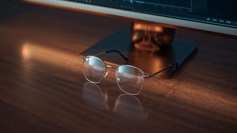 Metal-framed glasses on wooden desk with monitor reflection