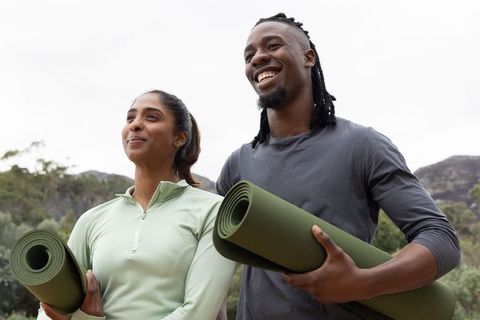 Diverse couple carrying rolled green yoga mats outdoors, smiling and preparing for nature yoga sessi