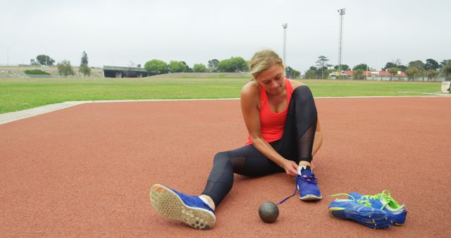 A female athlete in athletic wear tying her shoelaces on an outdoor track field. She is preparing for a sports activity, reflecting focus and determination. Ideal for use in fitness blogs, sportswear advertisements, motivation posters, or health and wellness articles.