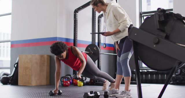 Image of diverse female fitness trainer instructing woman raising dumbbells working out at a gym. Exercise, fitness and healthy lifestyle.